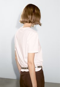 Light pink cotton t-shirt with short sleeves, paired with dark brown pants and a silver square-buckled belt. Smooth texture, minimal design.