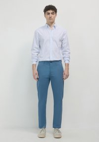 Mann in hellblauem, gestreiftem Hemd mit Knöpfen, blauen Hosen und beigen Schnürschuhen, der vor einem hellen, einfarbigen Hintergrund steht.