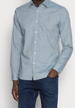 Hombre con camisa azul clara abotonada con pequeño logo bordado en el pecho y vaqueros azul oscuro, mano en el bolsillo.