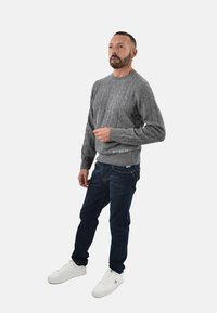 Pullover grigio a maglia rasata con polsini e orlo a coste, abbinato a jeans blu scuro e sneakers bianche. Dettaglio del logo discreto all'orlo.