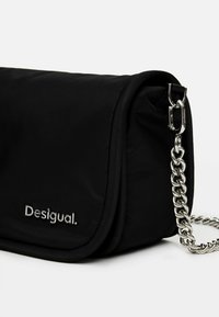 Czarna torba na ramię z miękkim materiałem zewnętrznym, klapką na złożenie, srebrnym paskiem na łańcuszku oraz metalowym logo "Desigual.".