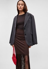 Grijze oversized blazer over een aansluitende bruine jurk met een franjerand. Rode handtas in de hand, tegen een neutrale achtergrond.