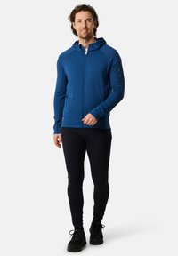 Felpa blu con zip e design aderente, dotata di cappuccio e tasche laterali, abbinata a leggings neri e sneakers nere.