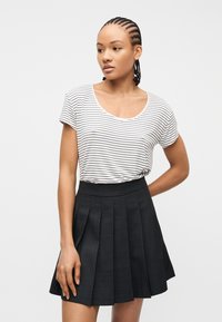 Γυναίκα με πλεκτά μαλλιά, φορώντας λευκό και μαύρο ριγέ πουκάμισο με κοντά μανίκια, που είναι μέσα σε μαύρη μίντι φούστα με πιέτες, στέκεται μπροστά σε λευκό φόντο.