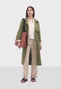 Abrigo largo verde oliva, blusa beige, pantalones crema y sandalias negras con estampado. Bolso rojo con diseño lineal blanco. Textura de tela suave.