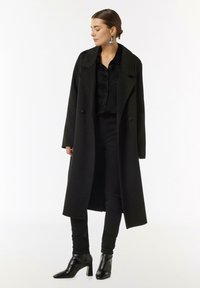 Black wool coat with a double-breasted design, wide collar, and long sleeves. Paired with a black satin shirt, skinny pants, and heeled boots.