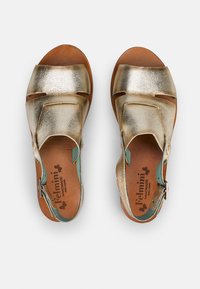 Sandales métalliques dorées avec deux larges bandes croisées, une boucle réglable et des accents teal contrastants sur le talon. Semelle intérieure en cuir.