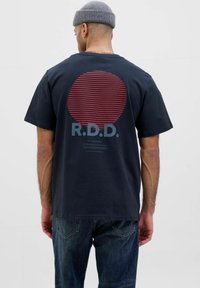 Tums zila kokvilnas T-kreklu ar sarkanu svītru apli grafikā uz muguras. Iezīmē "R.D.D." tekstu gaiši zilā krāsā un brīvu piegriezumu.