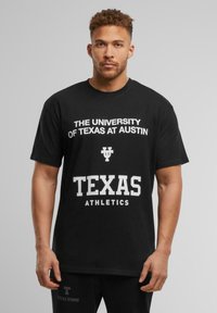Sort bomulds T-shirt med hvid tekst: "THE UNIVERSITY OF TEXAS AT AUSTIN" og "TEXAS ATHLETICS" i fede, blokbogstaver.