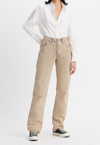 Jean beige taille haute avec une coupe droite, texture décolorée et détails de couture subtils, associé à une chemise blanche à boutons et des baskets noires.