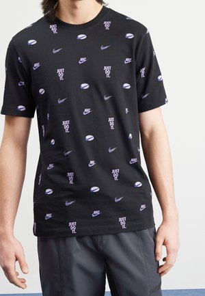 Hombre vistiendo una camiseta negra con logotipos púrpuras repetidos de Nike y el texto "Just Do It." combinada con pantalones gris oscuro.