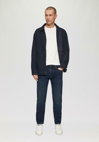 Veste bleu marine avec fermeture à boutons sur un t-shirt blanc, associée à un jean foncé et des baskets blanches. Design minimaliste, coupe décontractée.