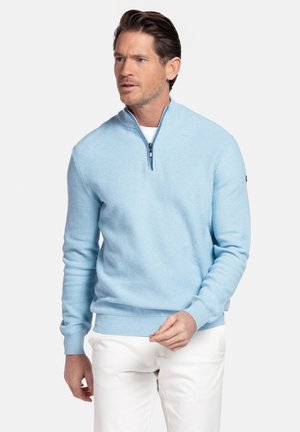 Man met lichtblauwe trui met kwartrits en witte broek, kijkt lichtjes opzij met een neutrale gezichtsuitdrukking.