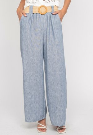 Femme portant un pantalon large bleu clair avec une ceinture beige et des sandales blanches à lanières, mains dans les poches, debout sur un fond blanc.