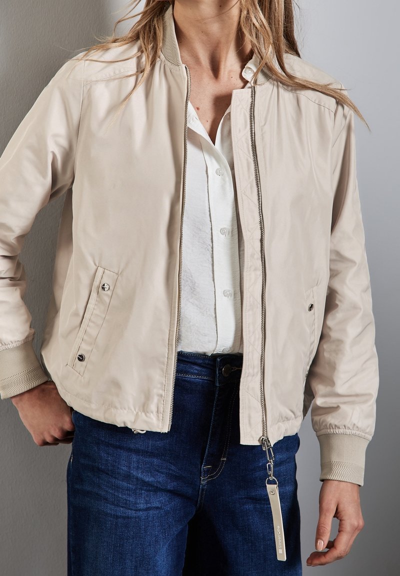 Femme portant une veste bomber beige sur une chemise blanche à boutons et un jean bleu, une main reposant près de la taille.