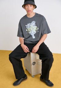 T-shirt gris surdimensionné avec un imprimé graphique audacieux jaune et blanc, associé à un pantalon noir et des chaussures noires, assis sur un conteneur argenté.