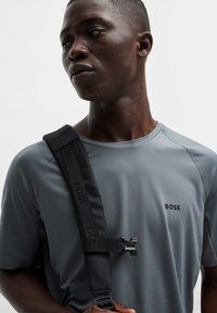 T-shirt de sport gris à texture lisse, avec un logo noir sur la poitrine et une sangle noire avec une boucle sur l'épaule.