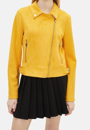 Femme portant une veste zippée jaune vif avec des boucles latérales et une jupe plissée noire.