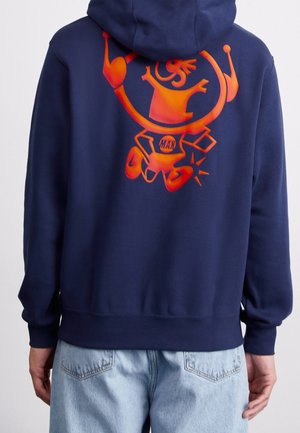 Person iført en mørkeblå hættetrøje med en lys orange abstrakt tegneseriefigur og ordet "MAX" på bagsiden, kombineret med lyseblå jeans.