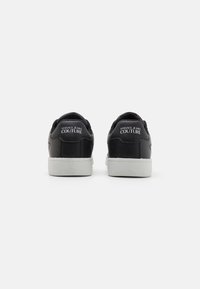 Svarta lädersneakers med vita gummisulor. Bakre vy visar "VERSACE JEANS COUTURE"-logotypen på hälen. Mjuk struktur och minimalistisk design.