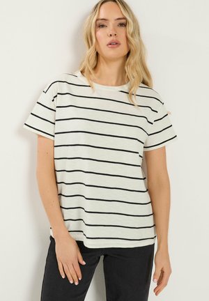 Pitkätukkainen vaaleahiuksinen nainen, joka pukeutunut valkoiseen, lyhythihaiseen paitaan, jossa on mustia vaakaraitoja, ja mustiin housuihin, seisoo valkoista taustaa vasten.