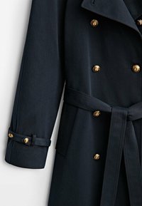 Manteau bleu marine foncé à double boutonnage avec boutons dorés, ceinture assortie nouée à la taille et lanière avec boutons dorés au poignet de la manche.