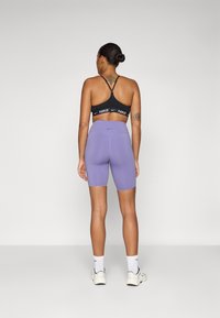 Soutien-gorge de sport noir avec logo blanc, associé à un short de vélo taille haute lavande. Texture lisse avec un design ajusté et un branding minimal.