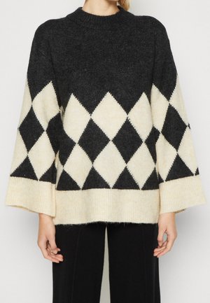 Pull en maille à motif losanges noir et blanc avec manches larges, porté avec un pantalon noir, visible de la moitié du torse jusqu'au cou.