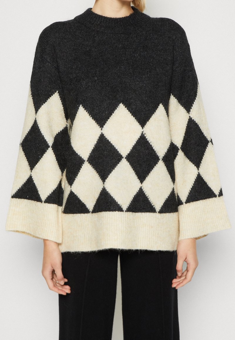 Pull en maille à motif losanges noir et blanc avec manches larges, porté avec un pantalon noir, visible de la moitié du torse jusqu'au cou.