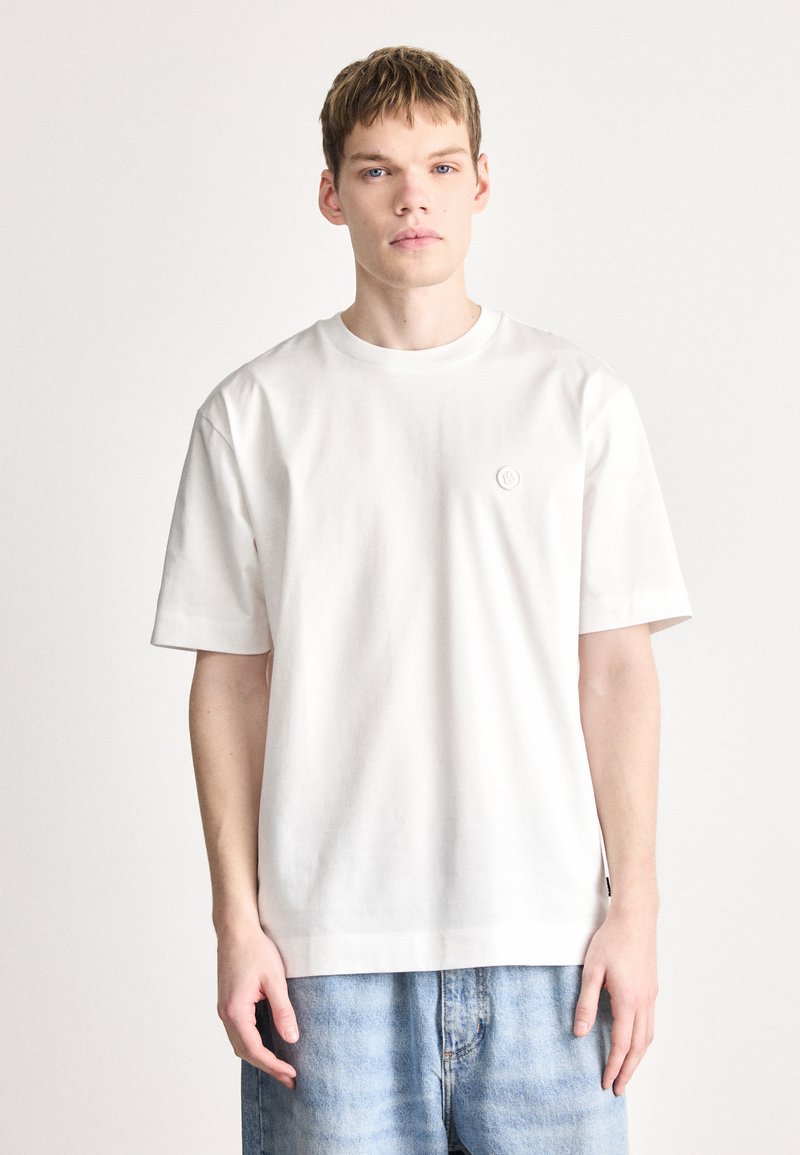 Tricou alb cu mâneci scurte, realizat din bumbac moale. Decolteu rotund, design simplu, cu un mic logo circular pe piept.