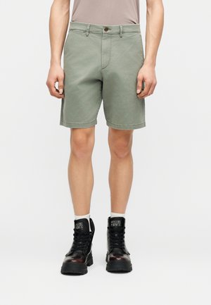 Άνδρας που φοράει ανοιχτό ελιάς πράσινο σορτς μήκους μέχρι το γόνατο, ανοιχτό μπεζ μπλουζάκι μπλουζάκι κρυμμένο μέσα, λευκές κάλτσες και μαύρα μποτάκια με κορδόνια.