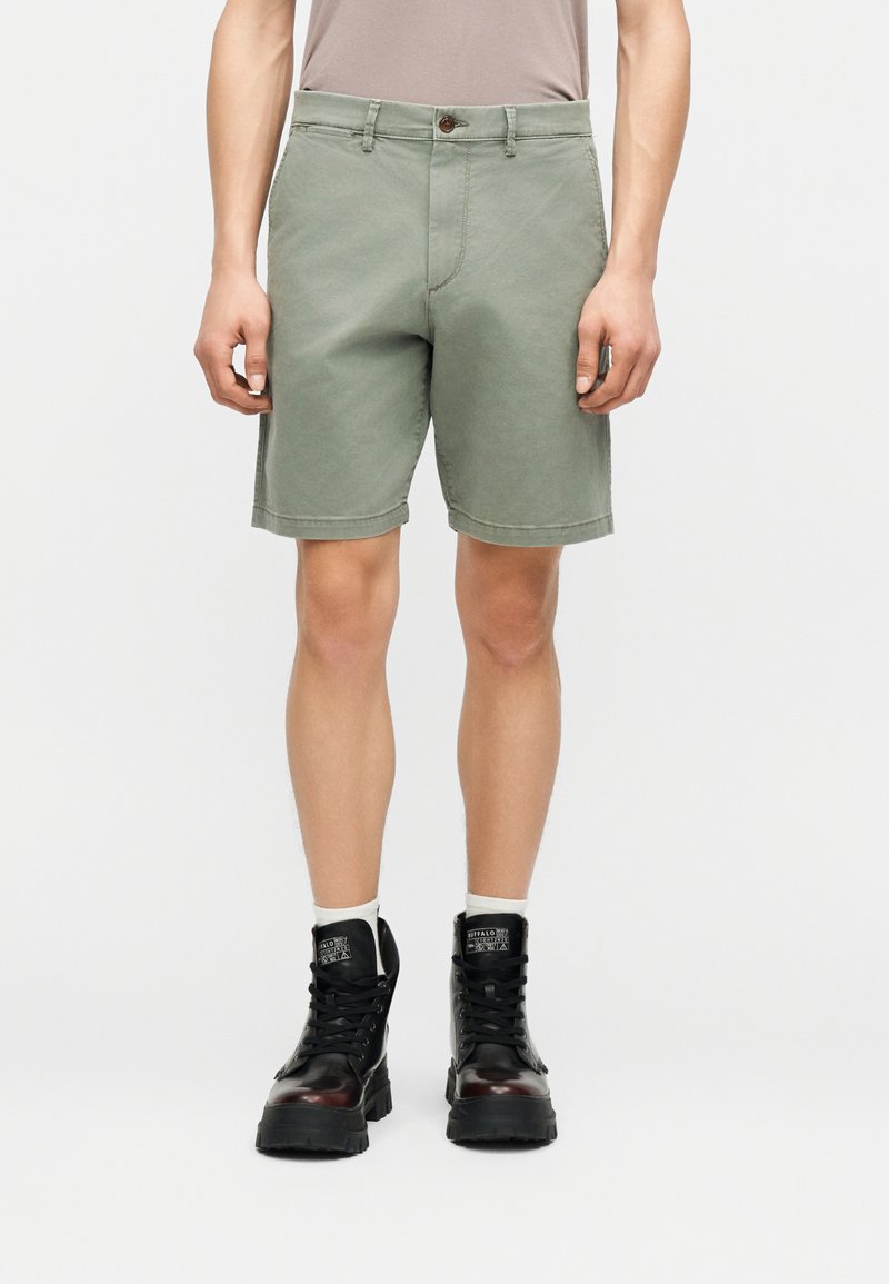 Άνδρας που φοράει ανοιχτό ελιάς πράσινο σορτς μήκους μέχρι το γόνατο, ανοιχτό μπεζ μπλουζάκι μπλουζάκι κρυμμένο μέσα, λευκές κάλτσες και μαύρα μποτάκια με κορδόνια.