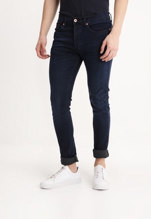 Jeans slim fit blu scuro arrotolati alle caviglie, indossati con sneakers bianche e top nero, mostrati dalla vita ai piedi.