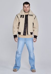 Beige shearling jacket with black leather accents and large collar, paired with light blue jeans. Features two chest pockets and ribbed cuffs.