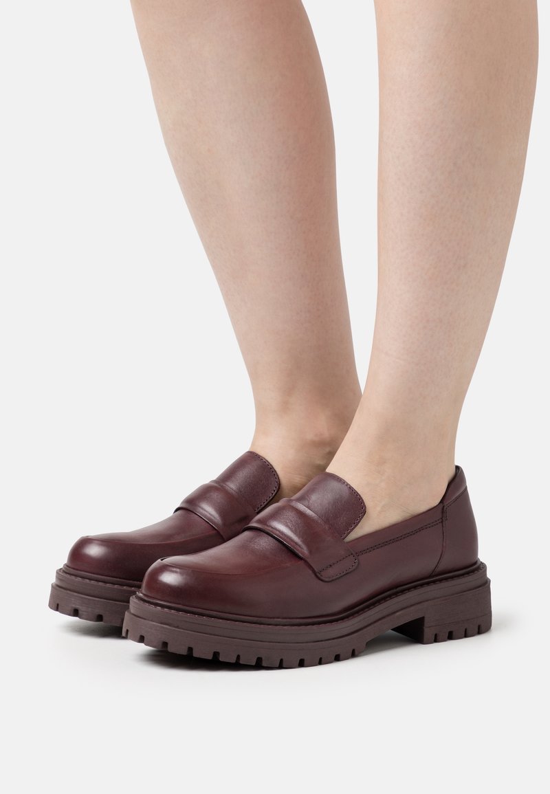 Mocassins épais en cuir marron foncé portés pieds nus, avec des semelles épaisses à rainures et une large bride sur le dessus.