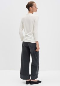 Vrouw met een knot, gekleed in een witte top met lange mouwen, zwarte wijde spijkerbroek met omslagen en zwarte ballerina's, staand met haar rug naar de kijker gekeerd.