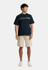 T-shirt in cotone blu navy con una striscia orizzontale beige e logo sul petto, abbinata a pantaloni corti beige e sneaker bianche.