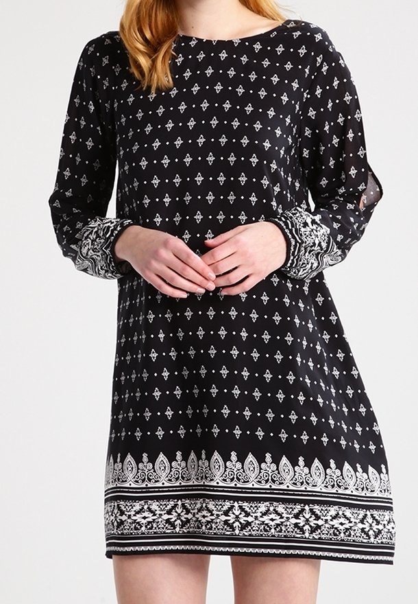 Femme portant une robe noire avec des motifs géométriques et floraux blancs, manches longues et poignets décoratifs, mains doucement jointes devant.
