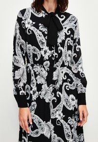 Vestido negro con un patrón floral y de cachemira en blanco. Tiene un cuello atado, mangas largas y una tela texturizada con un diseño fluido.