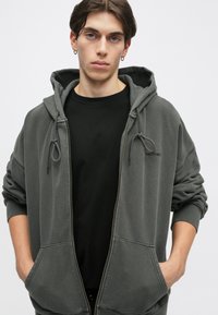 Hanorac gri cu fermoar și glugă ajustabilă, cu buzunare mari în față și manșete cu nervuri. Purtați peste un tricou negru, cu branding minim.