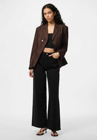 Blazer court marron avec un seul bouton doré, porté sur un top court noir, associé à un jean noir taille haute et large et des sandales noires.