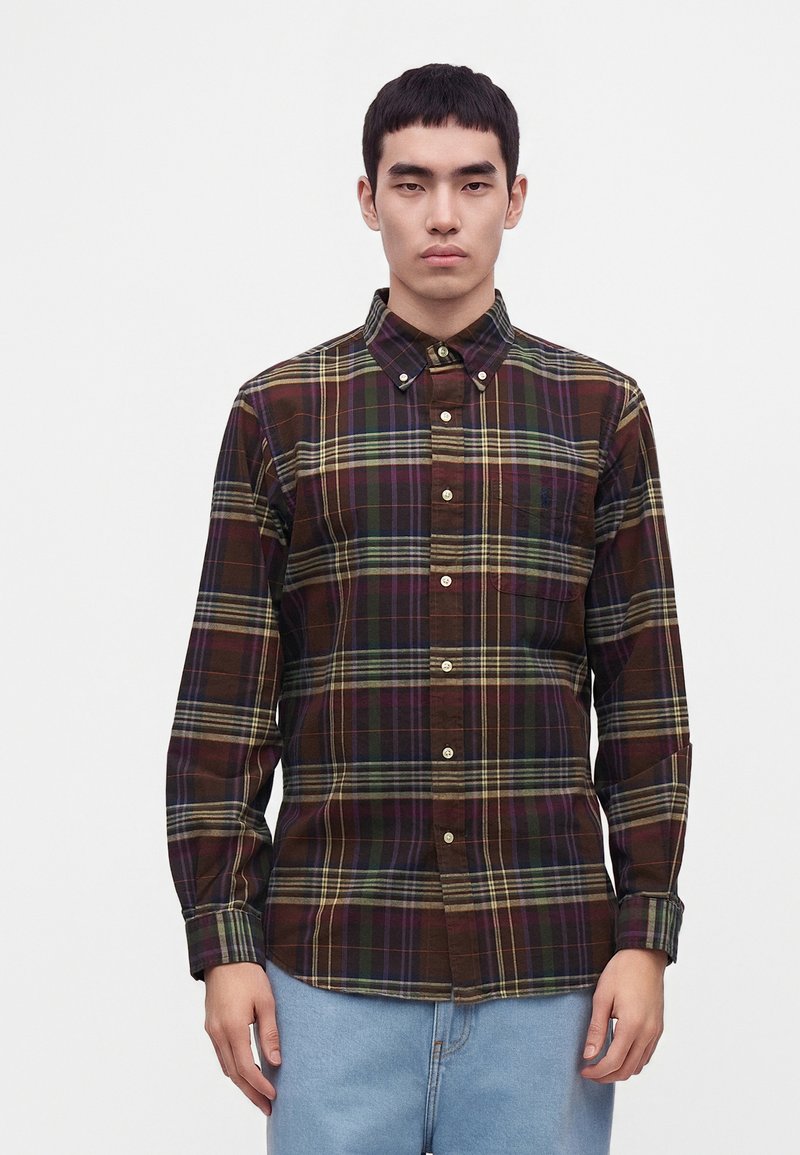 Homme portant une chemise à carreaux multicolores foncée à manches longues et un jean bleu clair, debout devant un fond blanc uni.