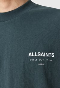 Chemise verte foncée avec le texte blanc "ALLSAINTS," des caractères japonais, et "LONDON" imprimé sur la poitrine près du col.