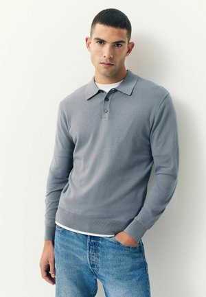 Graues langärmeliges Polo-Shirt mit flachem Kragen und zwei Knöpfen, kombiniert mit hellblauen Jeans. Sanfte Textur und taillierte Passform.