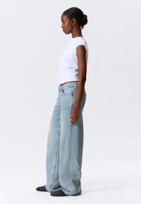Lichtblauwe, wijde spijkerbroek van denim, gecombineerd met een aansluitend wit t-shirt. Model staat zijwaarts, waarmee de relaxte pasvorm en silhouette worden getoond.