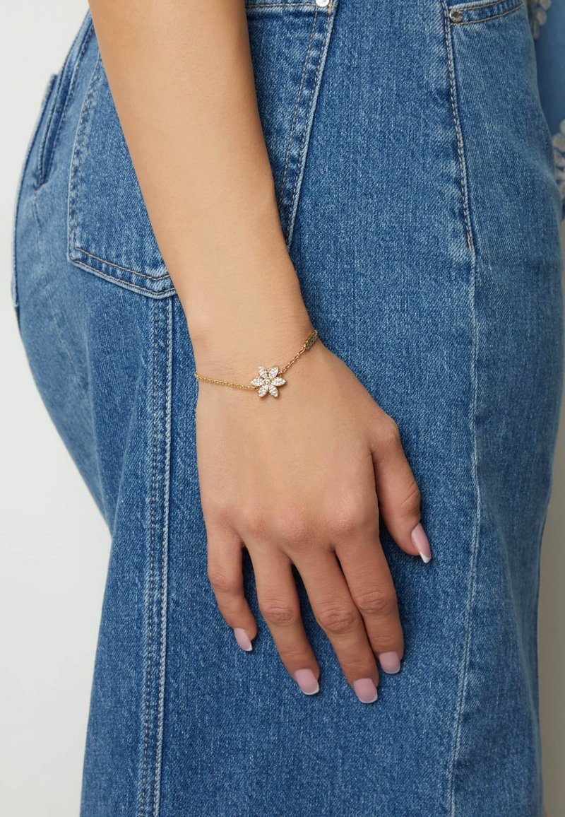 Gouden kettingarmband met een bloemencharme met witte edelsteenbloemblaadjes, gedragen op een hand tegen een denimachtergrond.