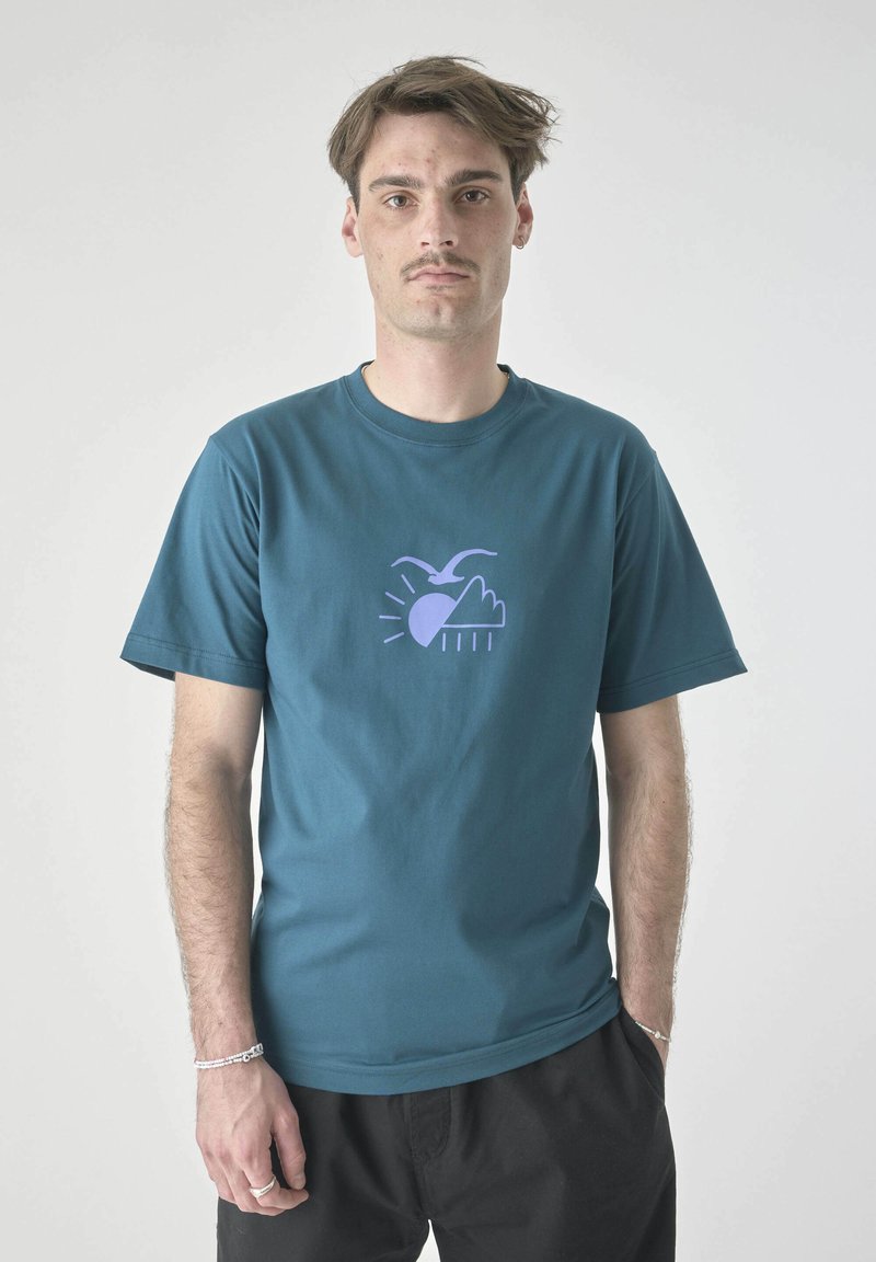 Teal Baumwoll-T-Shirt mit einem lila Grafikelement, das einen Vogel, die Sonne und eine Wolke mit Regen zeigt. Kurzärmlig, Rundhalsausschnitt, lockere Passform.