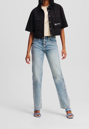 Personne portant un jean taille haute bleu clair avec braguette à boutons, une chemise noire courte boutonnée sur un haut beige, et des sandales bleues à lanières à franges.