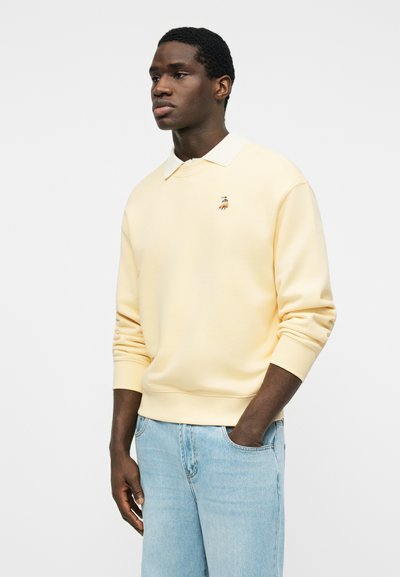 Νέος άνδρας που φοράει μια ανοιχτό κίτρινη φούτερ με ένα μικρό κεντημένο πάπια, συνδυασμένη με ανοιχτό μπλε τζιν, στέκεται μπροστά σε έναν απλό φόντο.