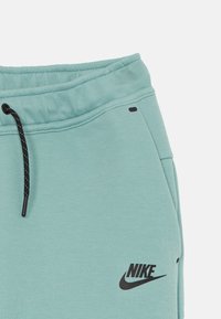 Gaiši mianti zaļi Nike sporta bikses ar nolaižamu jostu, melns logo un izšuvuma detaļām. Mīksta tekstūra un brīvs piegriezums.
